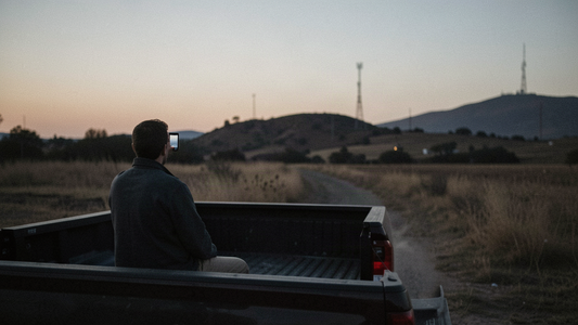 A person sitting on a pickup truck tailgate at dusk in a rural county, listening on a phone, with a radio tower on a distant ridge.